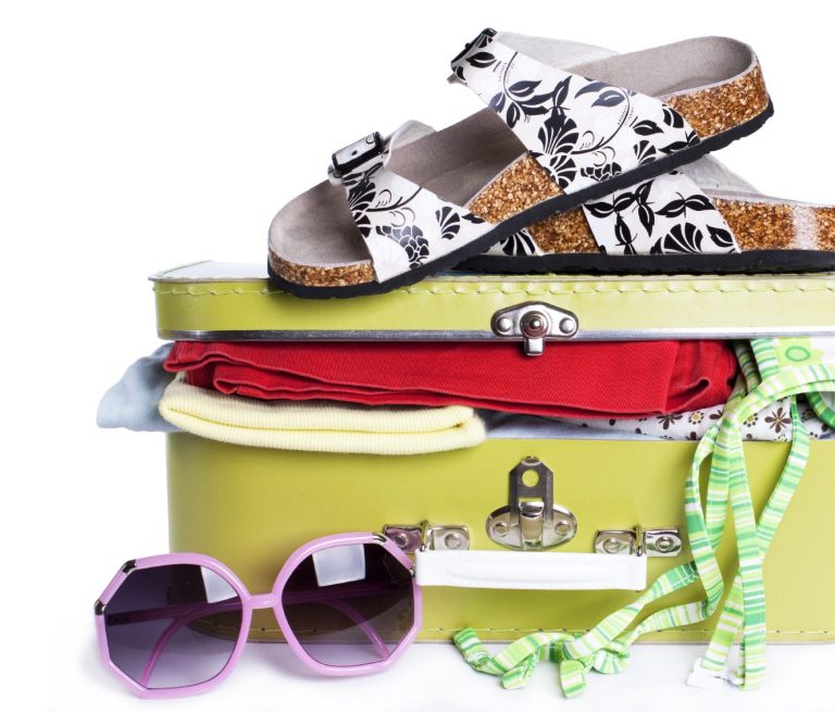 Schicke Sandalen, ein Koffer mit Kleidung und Sonnenbrille auf weißem Hintergrund.