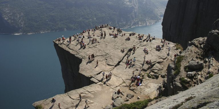 Felsformation mit Menschen auf einer Plattform über einem fjordartigen Gewässer.
