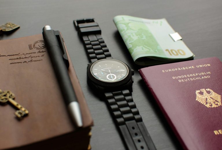 Schlüssel, Stift, Uhr, Geldschein und Reisepass auf einem Tisch angeordnet.