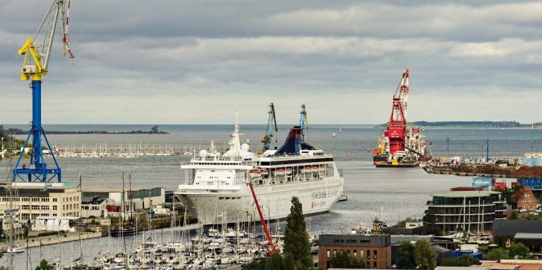 Hafenansicht mit Kreuzfahrtschiff und Krane im Hintergrund, bewölkter Himmel.