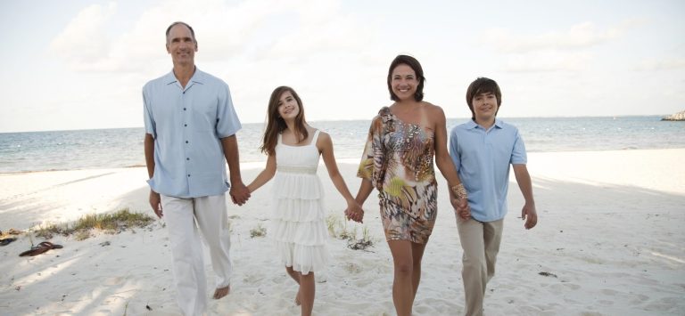 Familie geht Hand in Hand am Strand entlang, umgeben von Wasser und weitem Himmel.