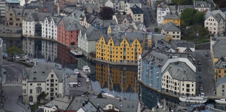 Bunte Häuser am Wasser mit Spiegelung, umgeben von Hügeln.